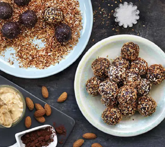 Dadelbollar recept: Mörka, kokosöverdragna energibollar på en ljusblå tallrik, bredvid en skål med mandelsmör och kakaopulver.