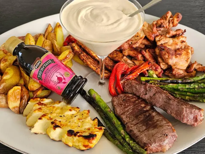 En festlig tallrik med grillad biff, kyckling, halloumi, potatis, sparris och en krämig sås. En flaska "Touch of Taste" sås till grillat står lutad mot potatisen.