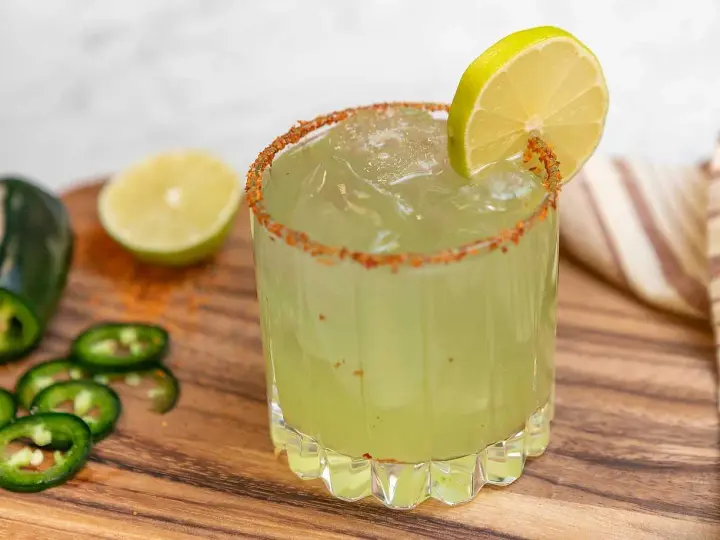 En glas med en kryddig margarita, garnerad med lime och chilipulver. Bredvid ligger jalapeños och limeklyftor.