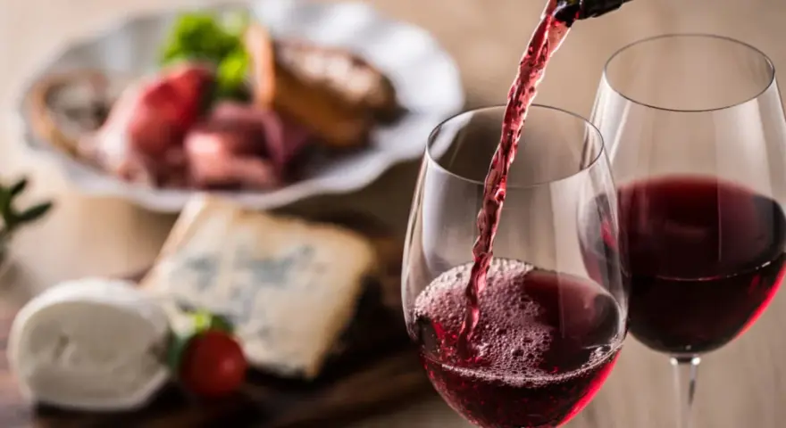 Rött vin hälls upp i glas, perfekt vin till grillat. Ost och charkuterier i bakgrunden.