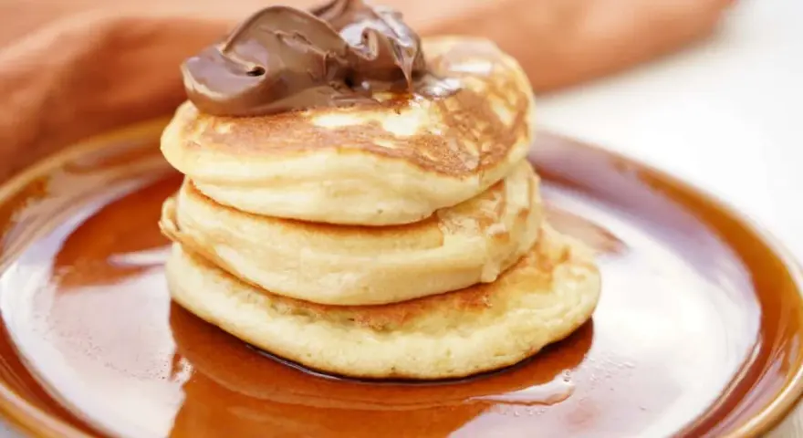 Stack of three fluffy American pancakes topped with chocolate spread on a brown plate.