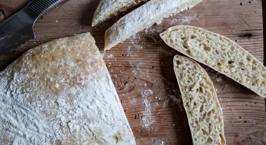 En hel och skivad ciabatta på en träskärbräda, redo för ett gott ciabatta recept. Mjöl strött över.