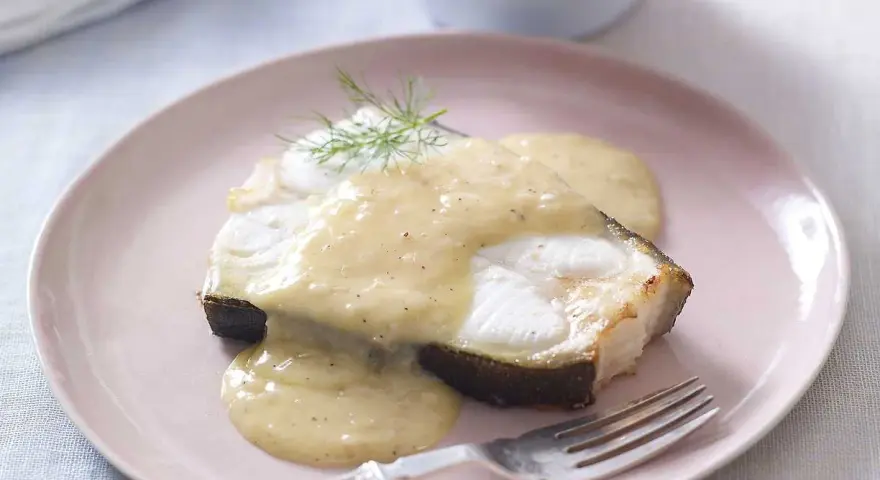En bit fisk täckt med krämig beurre blanc-sås, garnerad med dill, serveras på en ljusrosa tallrik med en gaffel.
