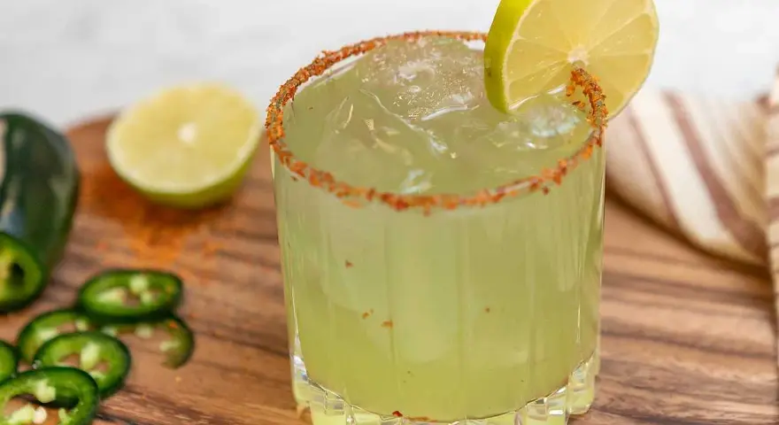 En glas med en kryddig margarita, garnerad med lime och chilipulver. Bredvid ligger jalapeños och limeklyftor.