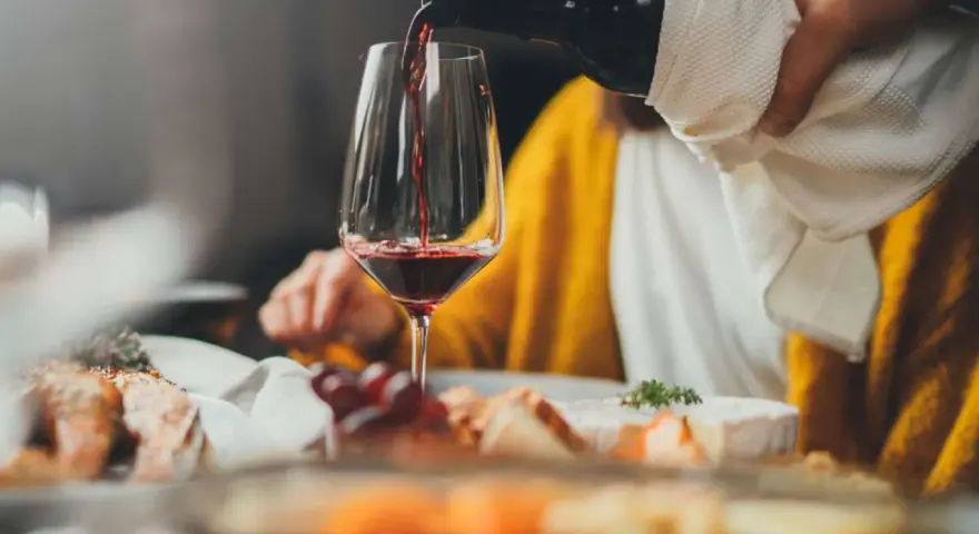 Rödvinet hälls upp i ett glas vid en middagsbjudning. En person i gul tröja sitter bakom. Frågan om hur mycket vin per person är relevant för en trevlig kväll.