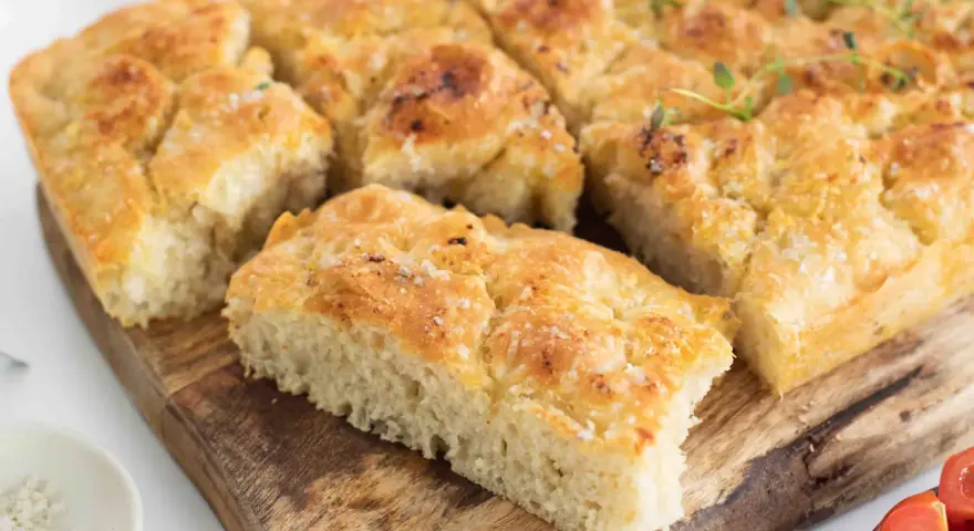 En gyllene focaccia långpanna, skuren i bitar, toppad med örter och flingsalt, ligger på en träskärbräda.