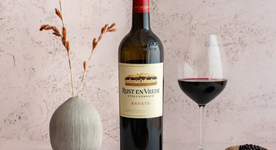 En flaska Rust en Vrede Estate vin, perfekt till oxfilé, står bredvid ett glas rött vin och en liten skål med pepparkorn.