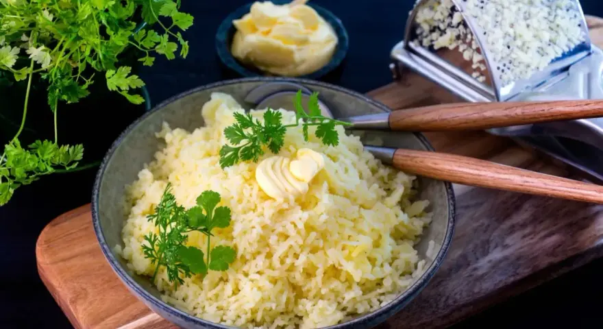 En skål med fluffig, pressad potatis toppad med smör och färska örter. Bredvid syns en smörskål och en rivjärn.