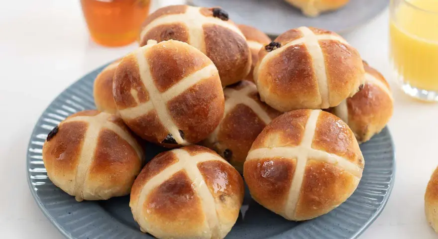 En hög med gyllene, nybakade hot cross buns på en blå tallrik, redo att avnjutas med honung och apelsinjuice.
