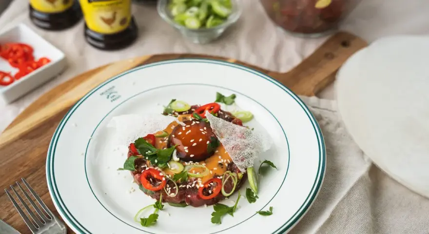 En läcker asiatisk råbiff med chili, salladslök och sesamfrön, serverad på en vit tallrik med gröna kanter.