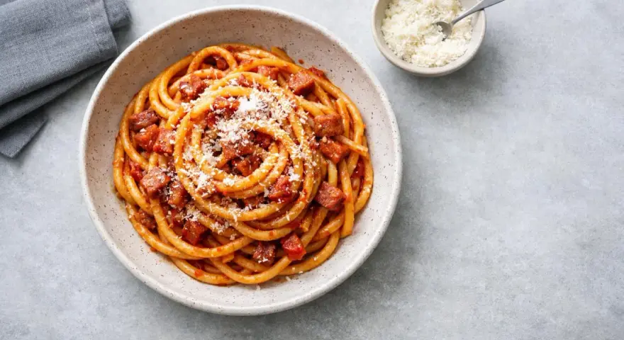 En skål med rörig pasta amatriciana, toppad med riven ost. En liten skål med mer ost och en grå servett syns bredvid.