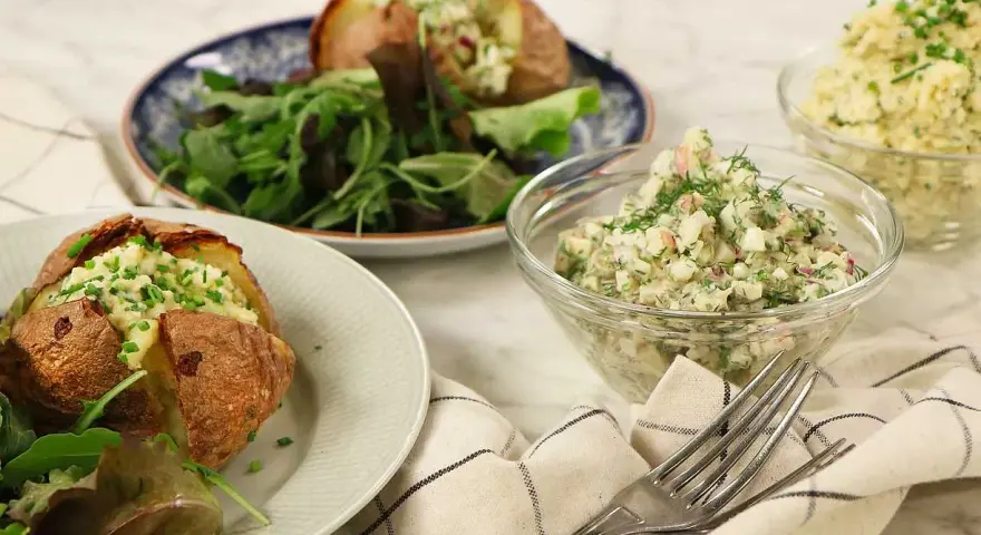 Två bakade potatisar med en krämig bakad potatis fyllning, serverade med en fräsch sallad och två skålar med olika röror.
