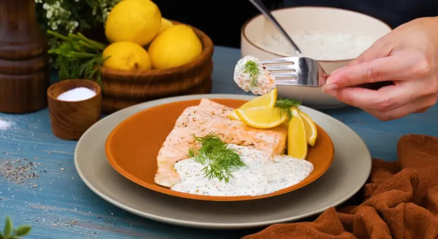En bit lax med citronskivor och dill, toppad med en krämig romsås till lax. En gaffel med romsås lyfts från skålen.