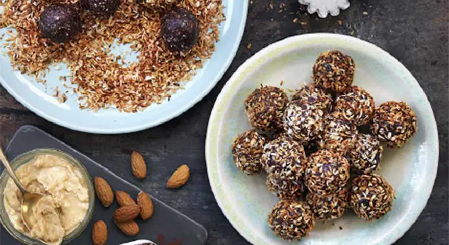 Dadelbollar recept: Mörka, kokosöverdragna energibollar på en ljusblå tallrik, bredvid en skål med mandelsmör och kakaopulver.