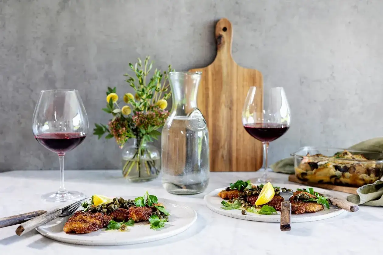 Två tallrikar med schnitzel, garnerade med kapris och citron, väntar på att avnjutas med ett glas rött vin till schnitzel. En vacker dukning.