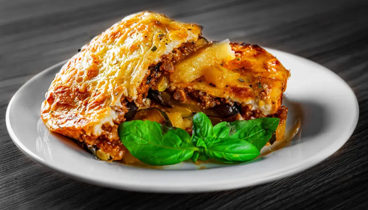 En bit gyllene moussaka med aubergine, köttfärs och béchamelsås, garnerad med färsk basilika. Ett utsökt moussaka recept.