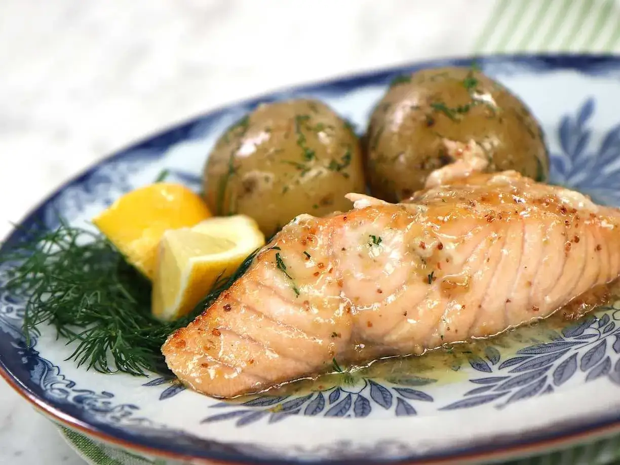 En saftig bit inkokt lax i ugn, serverad med dill, citronklyftor och små, örtkryddade potatisar på en vacker blåvit tallrik.