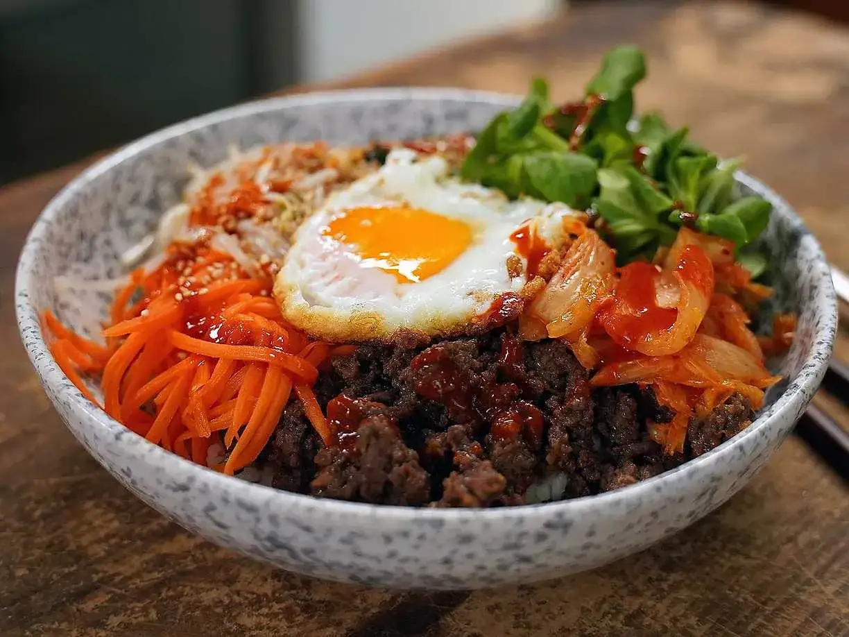 En skål med koreansk mat, bibimbap, med ris, köttfärs, strimlad morot, kimchi, en stekt ägg och grönsaker.
