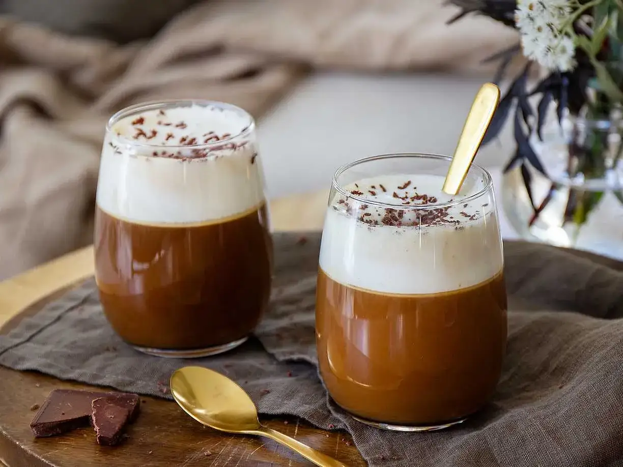 Två glas med krämig kaffe Karlsson recept, toppade med vispad grädde och chokladströssel. En guldig sked ligger i det ena glaset.