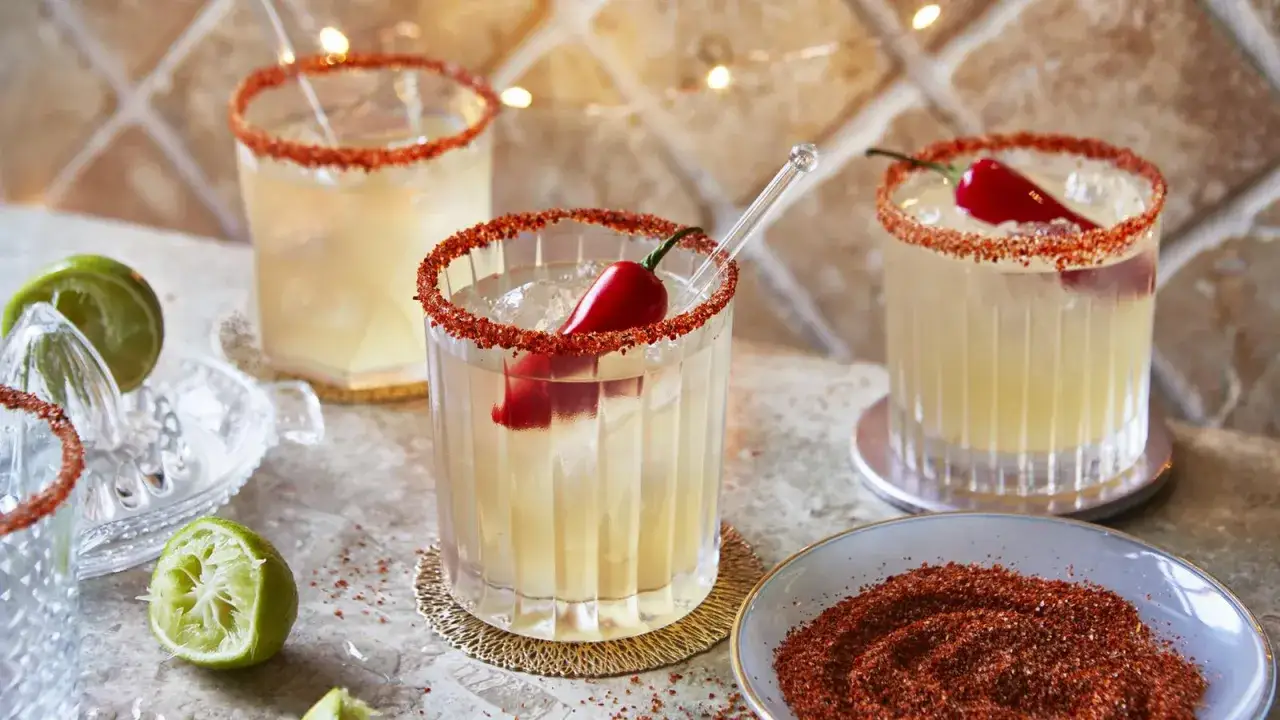 Tre glas med en kryddig margarita, dekorerade med chili och saltkant. Lime och en sk&aring;l med kryddblandning syns ocks&aring;.