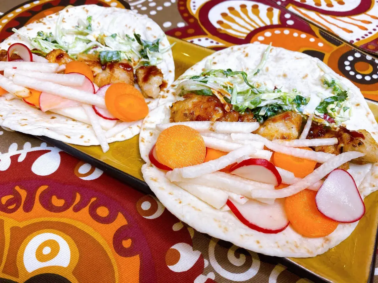 Två läckra asiatiska tacos med kyckling, krispig kål, rädisor och morötter på en färgglad duk.