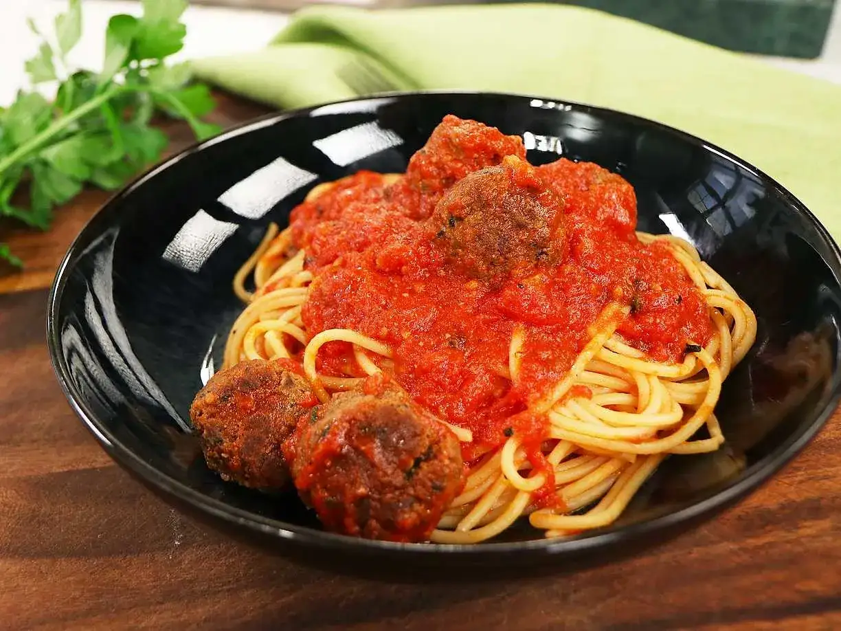 En skål med spaghetti och polpette al sugo, täckt med en rik tomatsås. Persilja och en grön duk i bakgrunden.