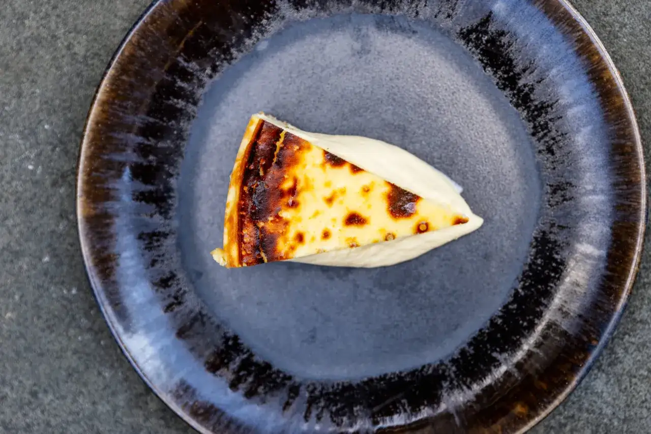 En bit baskisk cheesecake med karamelliserad yta, serverad p&aring; en m&ouml;rk tallrik med en kr&auml;mig s&aring;s.