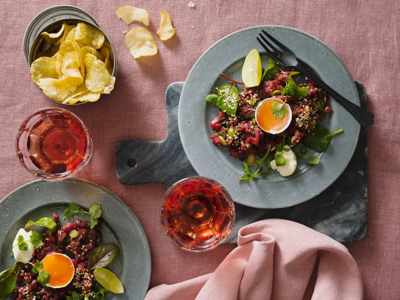 Två tallrikar med asiatisk råbiff, toppad med äggula och sesamfrön, serveras med rosévinsglas och potatischips.