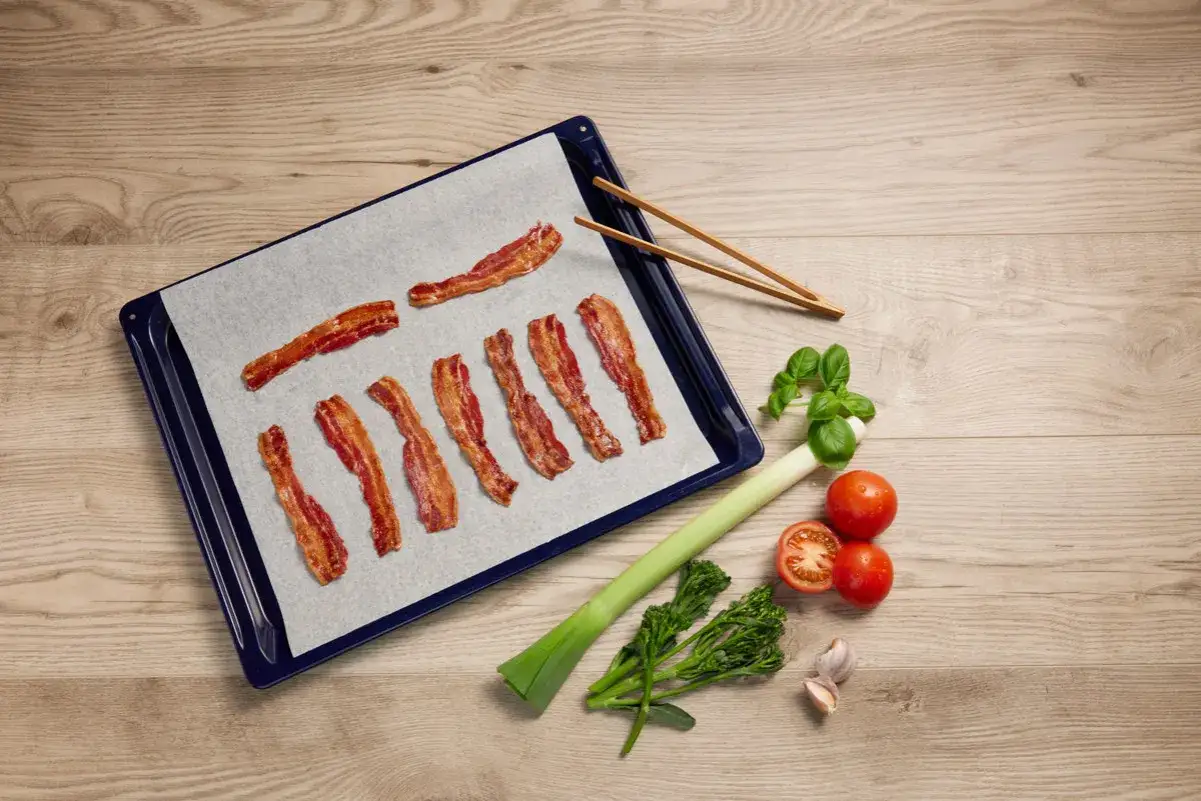 Krispigt bacon i ugn på bakplåtspapper, bredvid ligger grönsaker och vitlök.