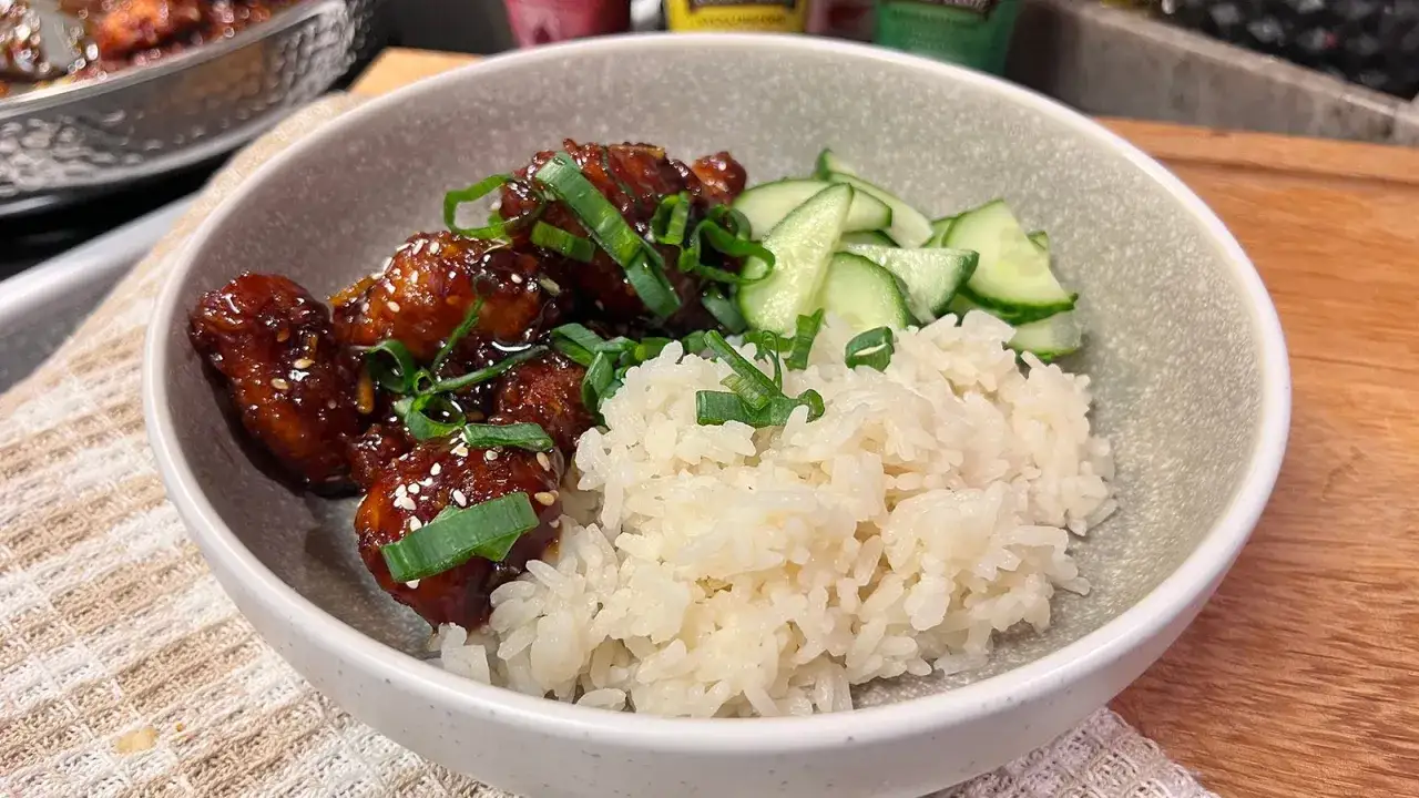 En skål med ris, skivad gurka och krispiga, glaserade bitar av sticky chicken, toppad med salladslök.