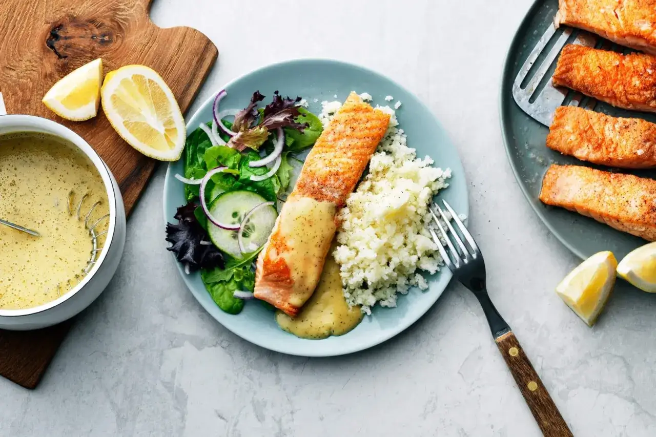 En tallrik med laxfilé, blomkålsmos och en fräsch sallad. En skål med krämig sås till laxen står bredvid, med citronklyftor som dekoration.