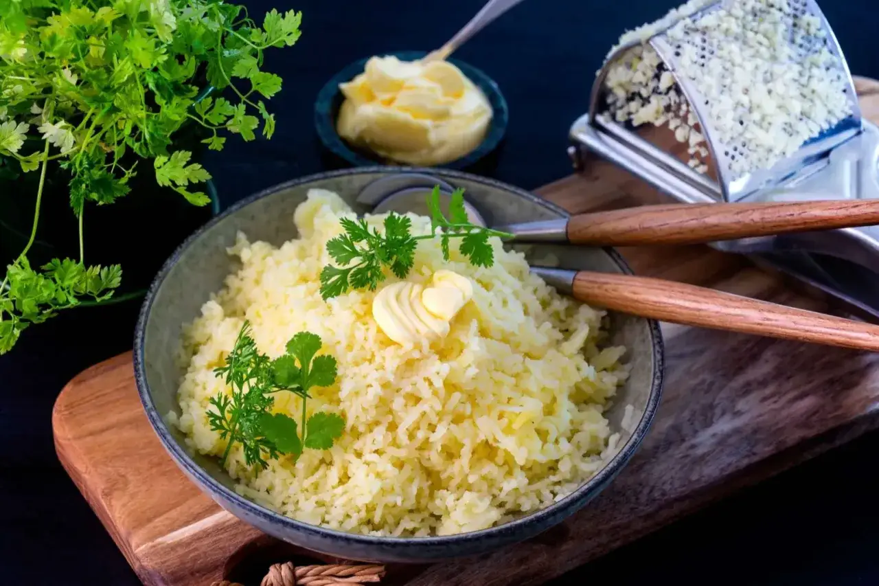 En skål med fluffig pressad potatis, toppad med en smörros och färska örter. Bredvid syns en smörskål och en rivare.