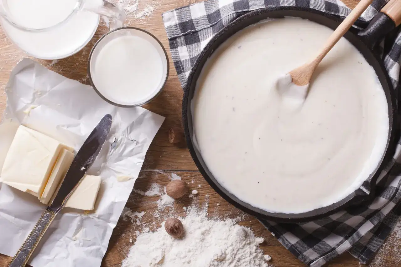 En kr&auml;mig bechamels&aring;s i en stekpanna, med ingredienser som sm&ouml;r, mj&ouml;lk, mj&ouml;l och muskotn&ouml;t redo f&ouml;r ett bechamels&aring;s recept.