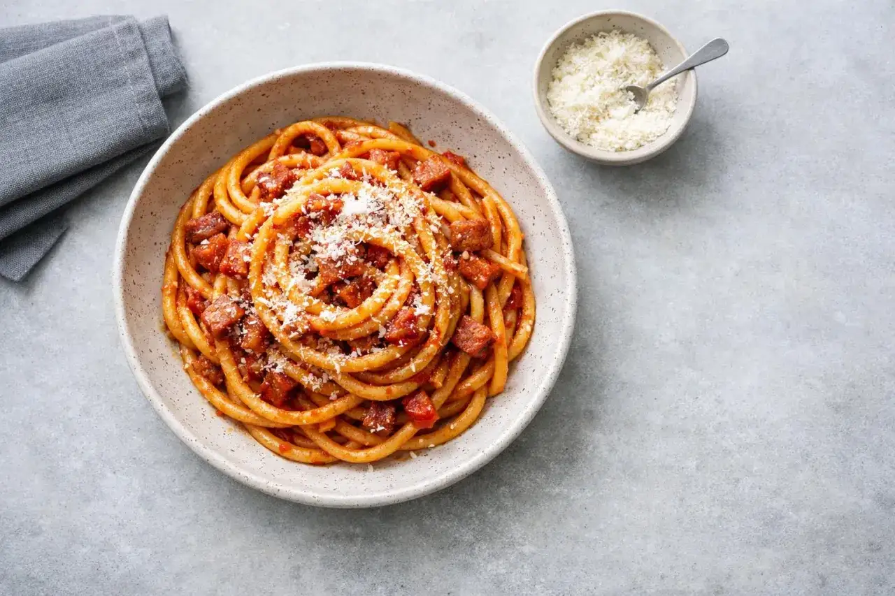 En skål med rörig pasta amatriciana, toppad med riven ost. En liten skål med mer ost och en grå servett syns bredvid.