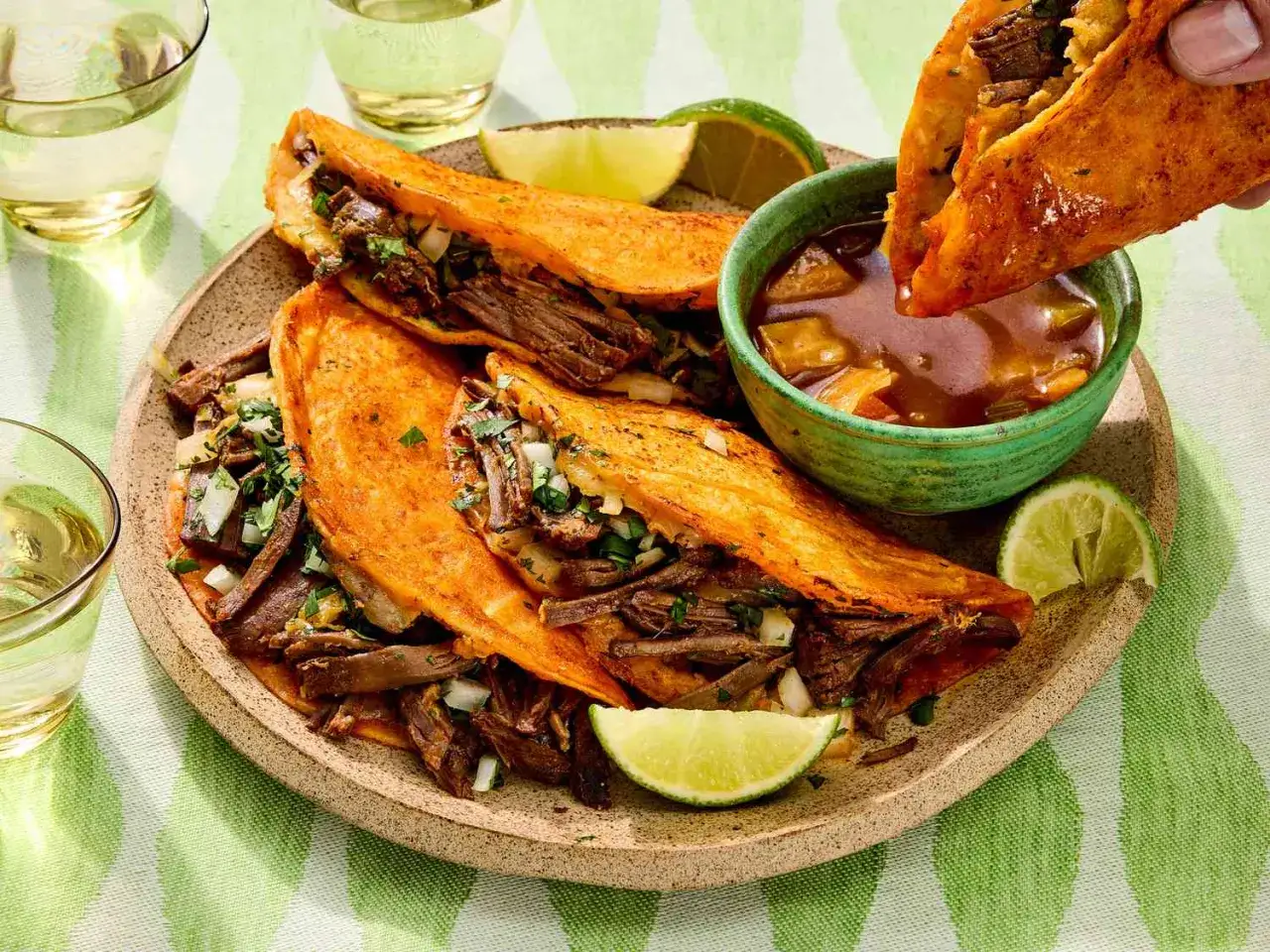 En hand doppar en krispig birria taco i en skål med buljong. Serveras med limeklyftor och mer tacos på en tallrik.
