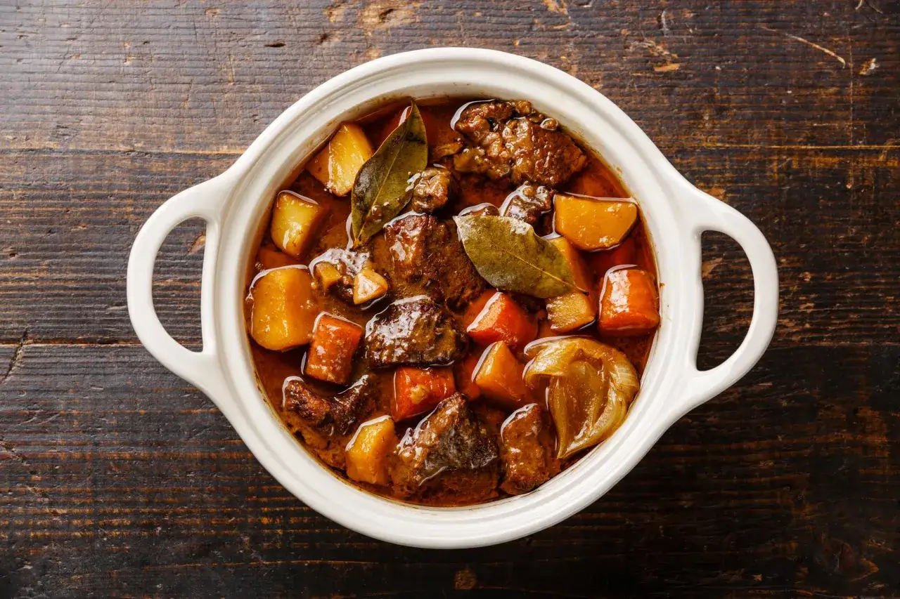 En vit keramikgryta fylld med mustig boeuf bourguignon, med möra köttbitar, potatis och morötter i en rik sås.