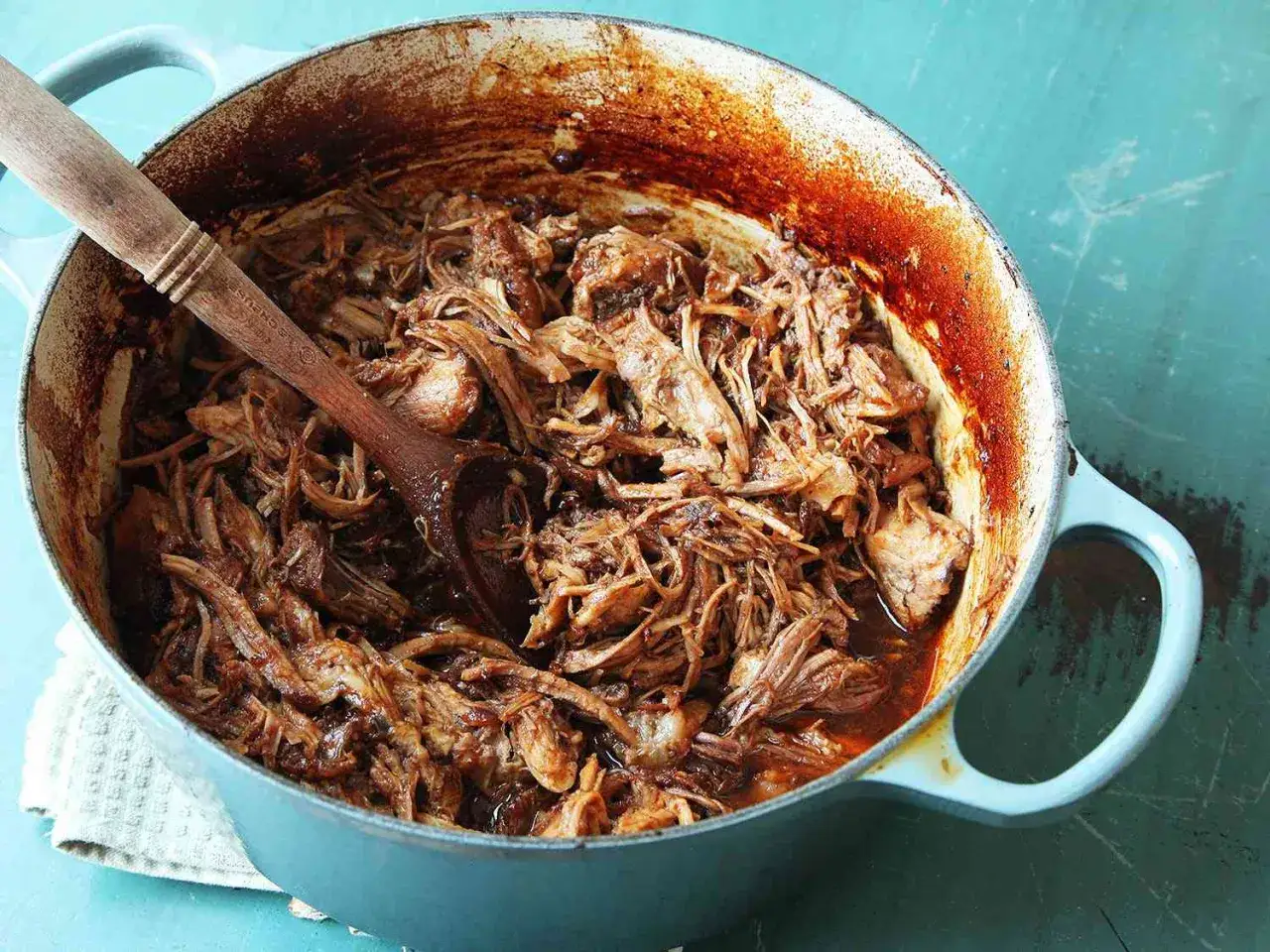 En stor gryta med saftig, mör pulled pork gryta. En träsked vilar i den rika, mörka såsen.