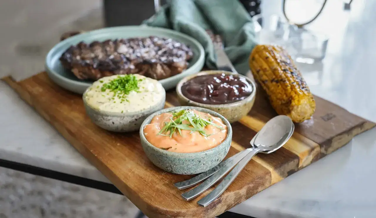 Grillad biff med majskolv och tre olika s&aring;ser till grillat: en kr&auml;mig vitl&ouml;kss&aring;s, en m&ouml;rk BBQ-s&aring;s och en rosa chilidressing.