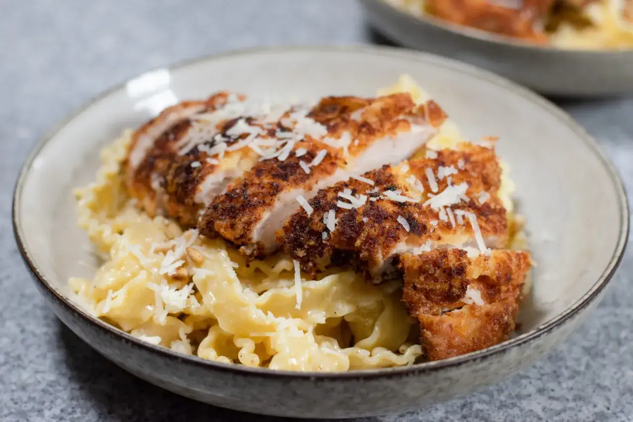 En skål med krusig pasta och krämig sås, toppad med saftig, skivad panerad kyckling och riven parmesan.