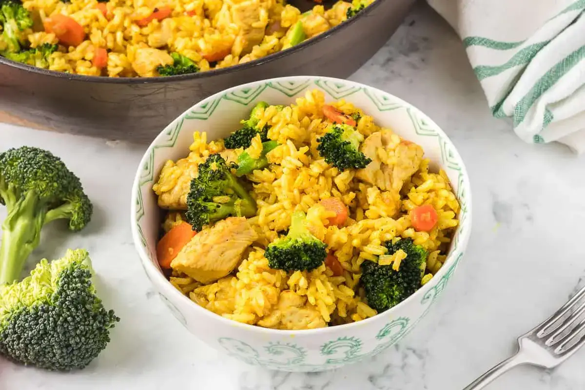 En skål med kyckling curry med ris, broccoli och morötter. En panna med samma maträtt syns i bakgrunden.
