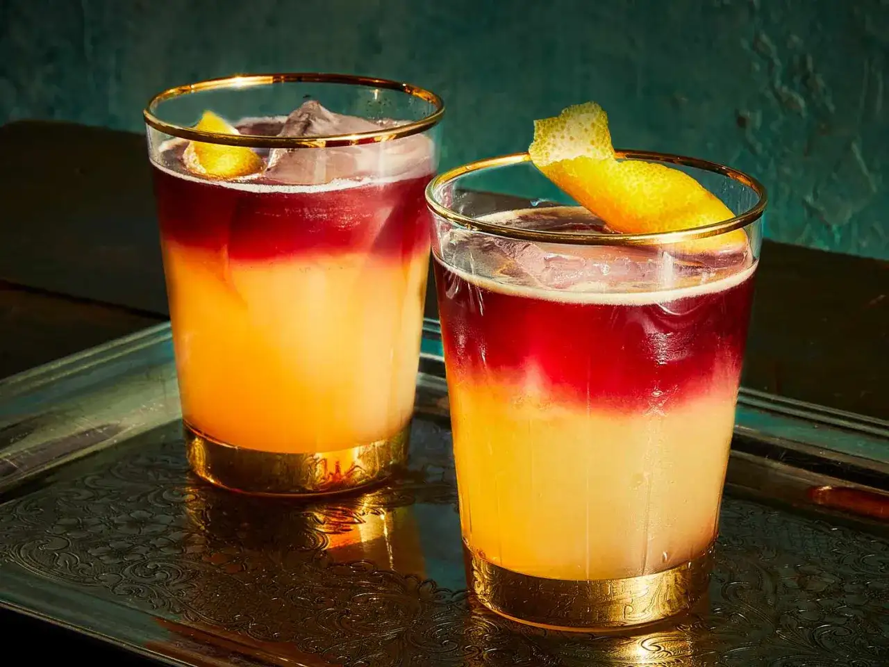 Två vackra New York Sour-drinkar med lager av orange och rött, garnerade med citronskal, serveras på en elegant bricka.