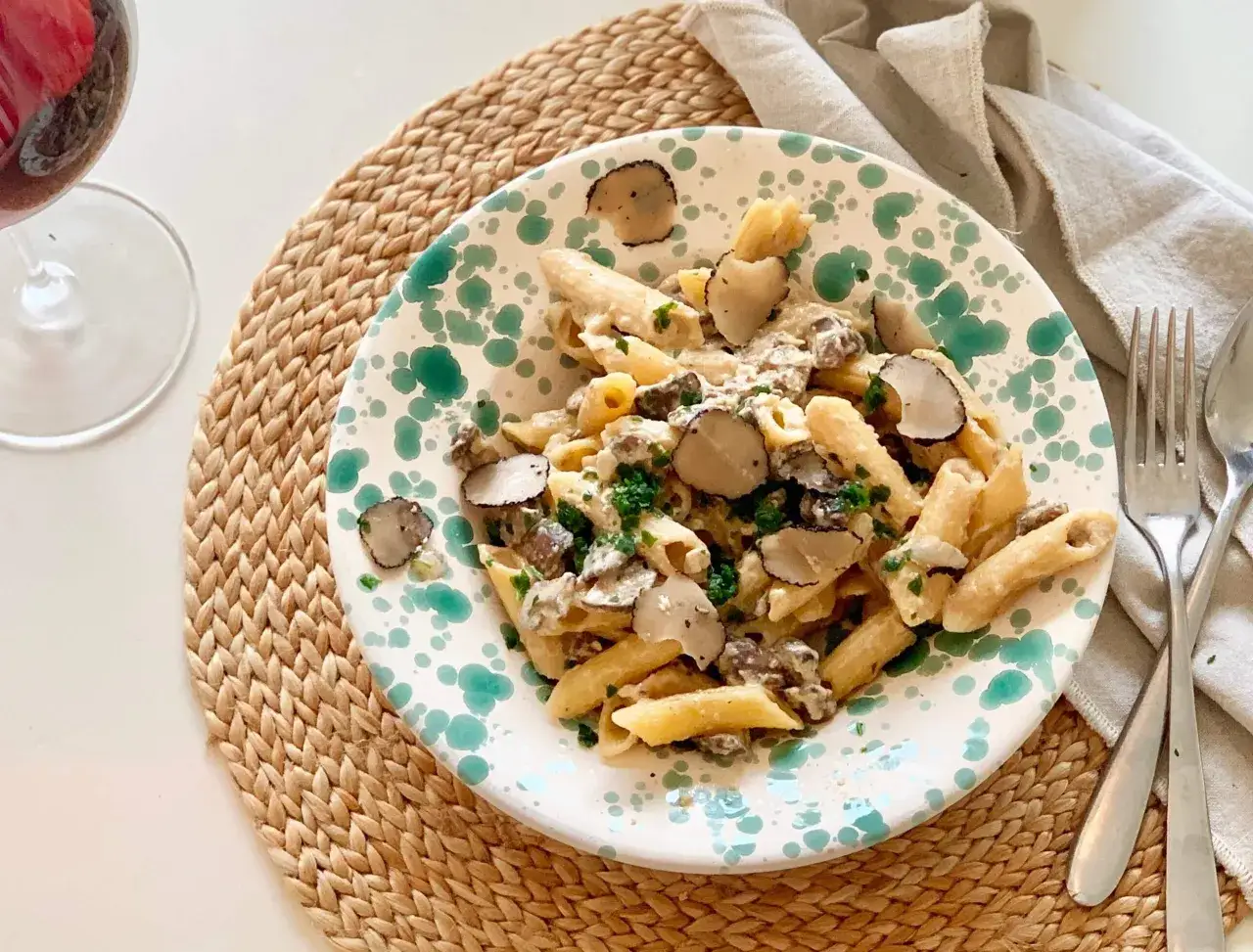 En tallrik med penne tryffelpasta, toppad med färsk tryffel och persilja. Ett glas rödvin står bredvid, perfekt för att ackompanjera denna lyxiga rätt.