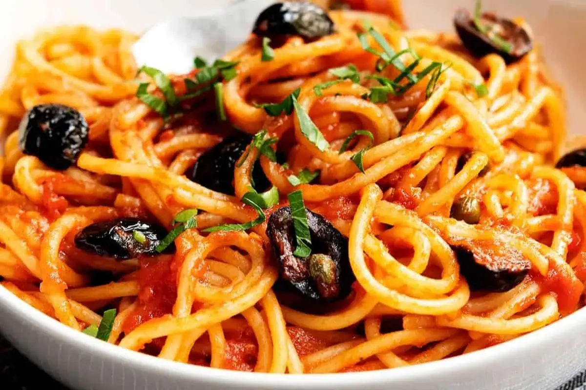 En skål med spaghetti puttanesca, toppad med svarta oliver och färska örter.