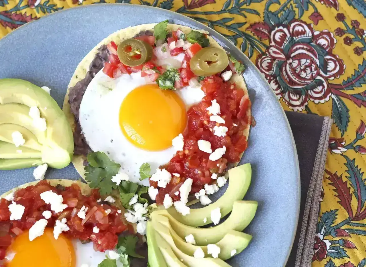 Två läckra huevos rancheros på en blå tallrik, med avokado, salsa, jalapeños och ost.