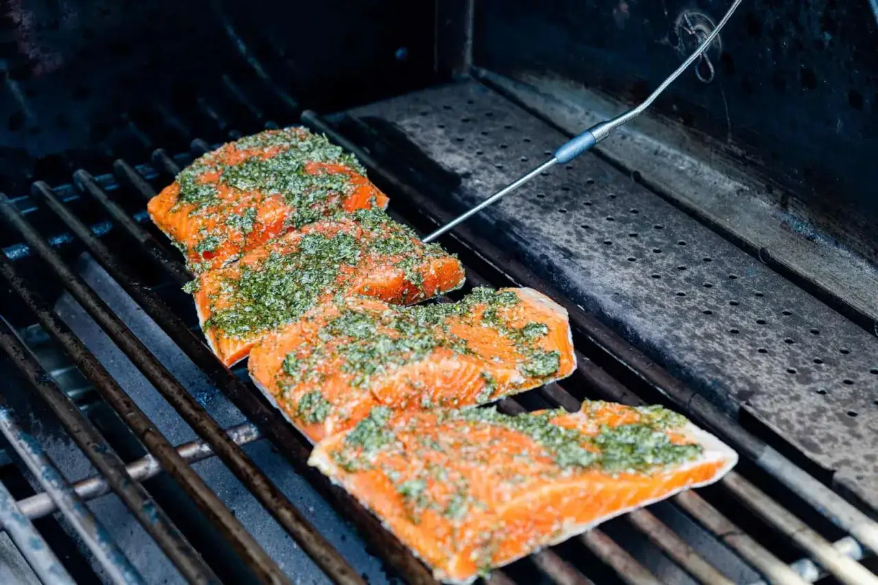 Tre saftiga laxfiléer med örtmarinad ligger på en gasolgrill. En termometer mäter temperaturen för perfekt grillad lax.