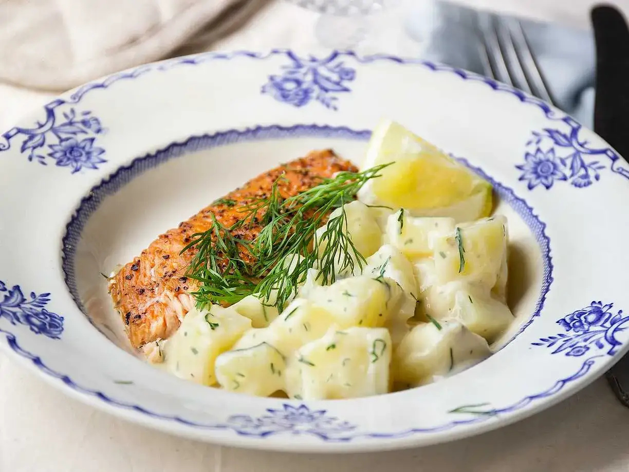 En bit lax med dill och en klyfta citron serveras med krämig stuvad potatis på en vacker blåvit tallrik.