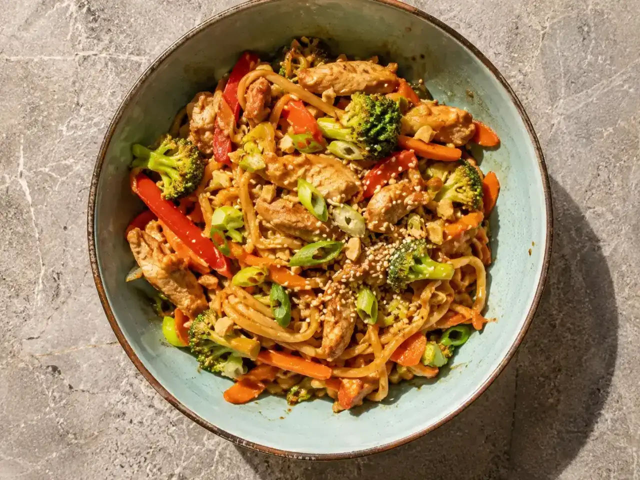 En skål med kycklingwok med nudlar, broccoli, morötter och paprika, toppad med sesamfrön.