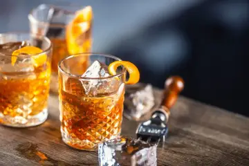 Tre old fashioned-drinkar med is och apelsinskal på ett träbord. En cocktailgaffel syns i bakgrunden.